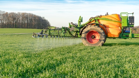 Landwirtschaftliche grüne Maschine mit orangem Tank sprüht mit Düsen Flüssigkeit auf darunterliegende Pflanzen unter blauem Himmel, im Hintergrund Baumzeile