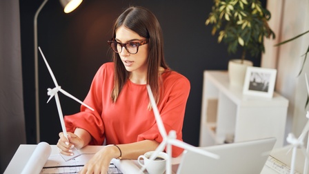 Person mit Brillen sitzt an Schreibtisch, deutet mit Finger auf Plan und hält in einer Hand Modell eines Windrades, ringsum weitere Windradmodelle und aufgeklappter Laptop