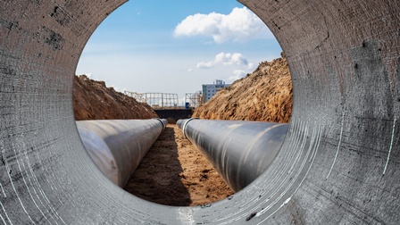 Ansicht aus einem Betonrohr das kreisrund Blick auf zwei schwarze Wasserrohe in Erdreich freigibt, links und rechts davon Erdwall, im Hintergrund verschwommen Konstruktionen einer Baustelle unter blauem Himmel