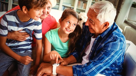 Zwei Erwachsene und zwei Kinder sitzen dicht nebeneinander auf einem hellen Sofa. Die Person ganz rechts hat am linken Handgelenk eine weiße Uhr mit einem Display. Eines der Kinder deutet mit dem Zeigefinger darauf. Alle blicken zur Person rechts