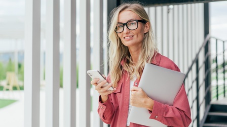Person mit langen blonden Haaren, Brille und rotem Hemd hält einen Laptop sowie ein Smartphone in Händen und blickt freudig in die Kamera während sie vor einem Hauseingang mit Lamellen steht