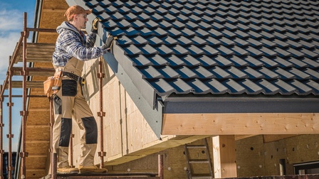 Eine Person mit Schutzbrille und breitem Gürtel mit Werkzeug steht auf einer erhöhten Plattform an einem Dach, bestehend aus dunklen Ziegeln. Die Person hält sich mit beiden Händen am Rand fest und blickt auf die Ziegel