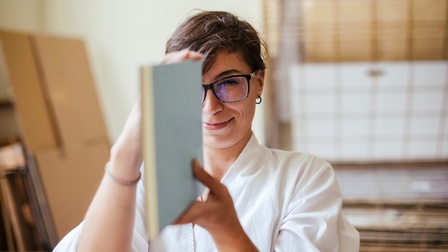 Eine Person mit Brille und weißem Hemd hält ein Buch mit hellblauem Umschlag vertikal vor ihren Kopf und blickt darauf.