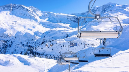 Bergige schneebedeckte Landschaft. Vereinzelt fahren Leute mit Skiern eine Piste hinunter. Eine Sesselliftbahn wird gekreuzt durch eine Gondelbahn. 