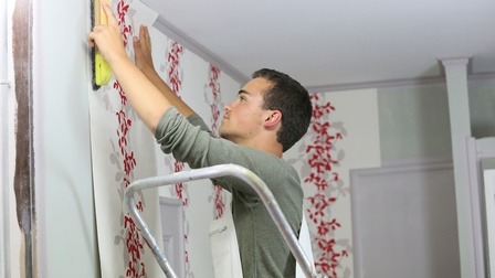 Person beim Anbringen von Tapeten an Wand, hält mit einer Hand das Tapetenpanel, stricht mit anderer Hand mit Bürste darüber