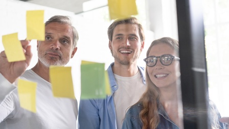 Drei Personen vor transparenter Tafel versammelt, auf der Post its kleben