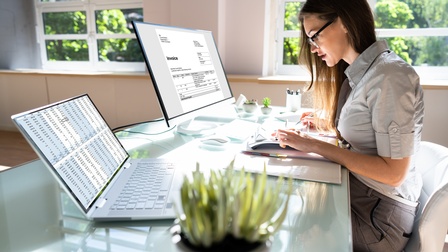 Person mit Brillen sichtet Unterlagen an Schreibtisch, vor ihr aufgeklappter Laptop und Computerbildschirm mit Tabellen und Rechnung