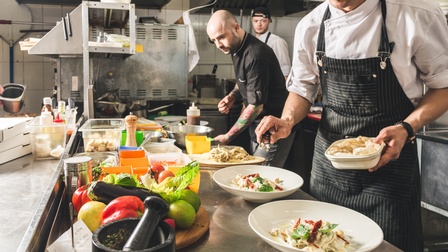 Detailansicht auf Teller in einer Gastronomieküche, ringsum kochende und Essen zubereitende Personen