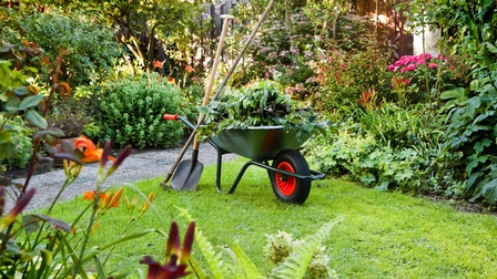 Schubkarre gefüllt mit Ästen und Blättern, daneben ist ein Spaten und ein Rechen angelehnt, im Hintergrund zeigt sich ein gepflegter Garten mit Sträuchern und Blumen