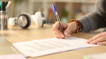 Auf einem Tisch liegen Papiere. Das oberste Blatt ist mit Text beschriftet. Die Hände einer Person liegen auf dem Blatt Papier. Die rechte Hand der Person hält einen Stift und schreibt damit etwas auf die untere Hälfte des Blattes