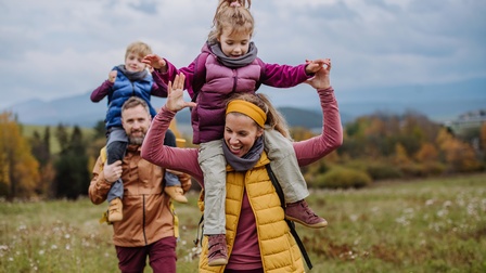 Zwei Erwachsene und zwei Kinder in Wanderkleidung gehen über eine Wiese. Je eine erwachsene Person trägt ein Kind auf ihren Schultern