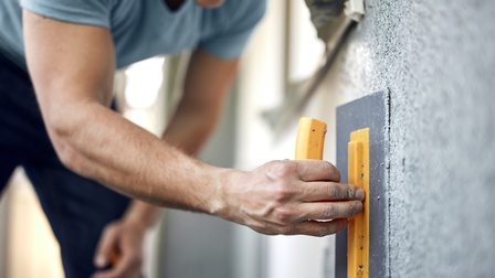 Nahaufnahme von einer Hand einer Person, die einen Putz auf einer Hausfassade mit einer Glättekelle aufträgt