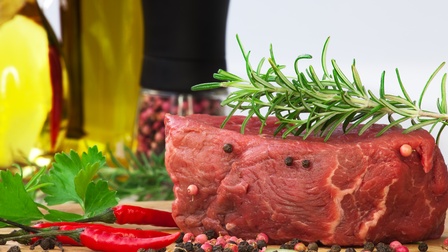 Rohes Stück Fleisch mit Rosmarinzweig darauf liegend, besetzt mit und ringsum verstreut Pfefferkörner, rote Chilischoten und Kräuter daneben liegend