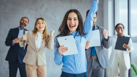 Lächelnde Person in Businesskleidung hält ein Tablet in einer Hand und streckt die andere Hand motiviert in die Höhe während weitere Personen freudig in einem modernen, hellen Raum hinter ihr stehen und in dieselbe Richtung blicken