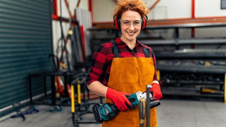 Portrait einer rothaarigen Person mit oranger Lederschürze, Schutzbrille, Gehörschutz und Handschuhen,  die Handschleifgerät in Händen hält in Werkhalle stehend, im Hintergrund verschwommen Regale