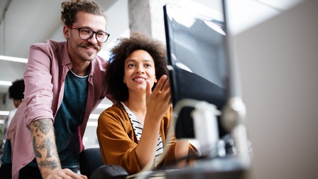 Eine Person sitzt bei einem Schreibtisch und blickt dabei auf einen Monitor, eine zweite Person mit Brille und Bart steht dahinter und blickt ebenfalls freudig auf den Monitor