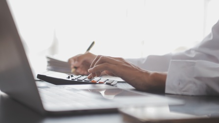 Auf einem Tisch steht ein aufgeklappter Laptop. Rechts daneben liegen Unterlagen und ein Taschenrechner. Auf den Tasten des Taschenrechners ist die linke Hand einer Person. Die rechte Hand hält einen Stift