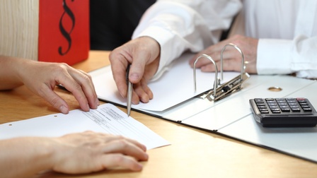 Zwei Personen, die eine Unterlage besprechen. Eine Person zeigt mit einem Stift auf eine Stelle im Dokument, daneben liegen ein Taschenrechner und eine Unterlagenmappe.