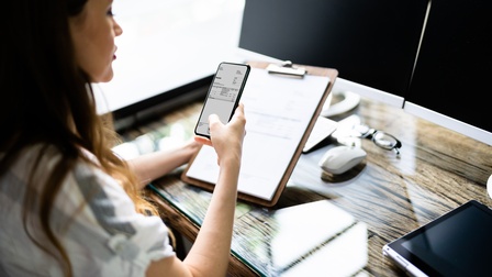 Person sitzt an einem Schreibtisch, hält ein Smartphone in der Hand und hat eine Rechnung geöffnet während gleichzeitig ein Klemmbrett gehalten wird, am Schreibtisch stehen Monitore, ein Tablet sowie eine Computermaus und eine Brille