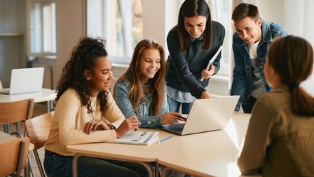 Mehrere lächelnde junge Personen um Tisch stehend und sitzend auf aufgeklapptes Notebook blickend und deutend