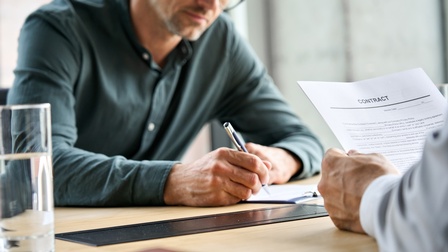 Person mit Brillen unterfertigt Dokument, Ausschnitt anderer Person, die Dokument mit Schriftzug Contract in Händen hält 