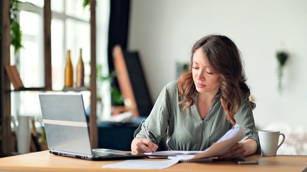 Eine Person sitzt an einem Tisch. Mit der rechten Hand notiert sie etwas in einem Heft. In der linken Hand hält sie Papiere. rechts vor ihr steht ein aufgeklappter Laptop.