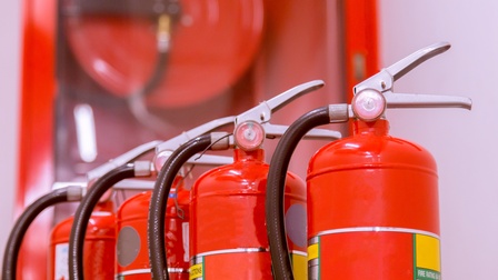 Vier zylinderförmige, rote Behälter stehen nebeneinander. Oben auf den Behältern sind jeweils zwei metallene Streben, die einen Schnabel bilden und an einen schwarzen Schlauch angeschlossen sind. Unten auf der Flasche steht dry chemical fire extinguisher