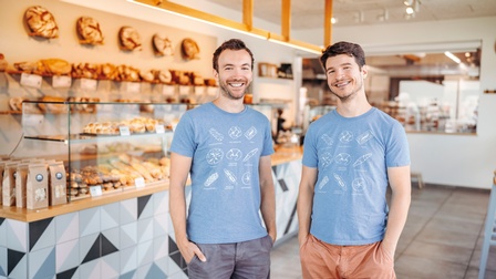 Zwei lächelnde Personen in blauen T-Shirt mit Motiven von Backwaren stehen in Bäckerei