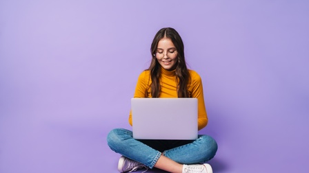 Jugendliche Person mit langen dunklen Haaren und Brille arbeitet im Schneidersitz mit einem Laptop vor einem lila Hintergrund