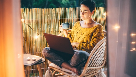 Person mit kurzen dunklen Haaren und gelbem Pullover sitzt mit einer Tasse und einem Laptop auf einem Balkon am Abend, dahinter erhellt eine Lichterkette die Atmosphäre
