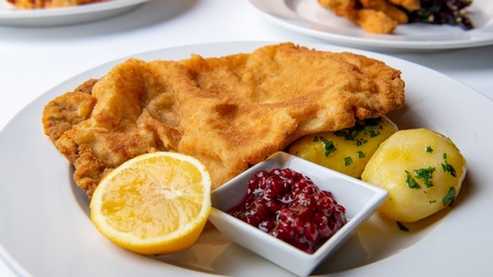 Nahaufnahme eines weißen Tellers, auf dem ein Wiener Schnitzel, Kartoffeln mit Petersilien, ein Schälchen mit Preiselbeeren und eine halbe Zitrone liegen.