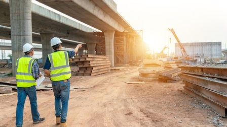 Rückenansicht zweier nebeneinander stehender Personen mit Helmen und neongelben Sicherheitswesten auf einem hellbraunen, erdigen Boden. Vor den Personen sind verschiedene Metallstangen, Betonteile und eine Brücke in Bauphase.