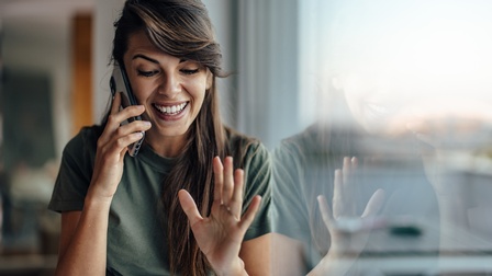 Person mit langen dunkelbraunen Haaren telefoniert und steht bei einer Fensterfron