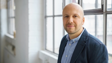 Portrait einer Person vor Fenster stehend mit blauem Hemd und Sakko 