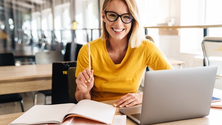 Lächelnde Person mit Brille und erhobenem Stift in der Hand sitzt vor aufgeklapptem Laptop und Büchern 