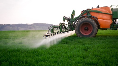 Orangegrüne landwirtschaftliche Feldspritzmaschine im Ausschnitt fährt über grün bewachsenen Acker und sprüht aus aus Düsen am Heck Flüssigkeit auf darunterliegenden Boden, im Hintergrund verschwommen begrünter Hügel unter grauem Himmel