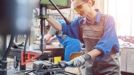 Person mit Schutzbrille, Schutzhandschuhen und in Arbeitsoverall bedient große Bohrmaschine und hält Zwinge mit eingeklemmtem Metallstück mit Hand, auf das sich Bohrkopf senkt