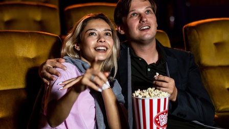 Frontalaufnahme zweier Personen, die nebeneinander in Kinosesseln sitzen. Die linke Person zeigt auf die Leinwand. Zwischen den Personen steht ein Kübel mit Popcorn. Die rechte Person hält Popcorn in den Händen