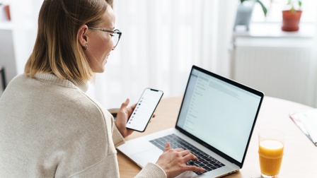 Lächelnde Person mit Brillen in Seitenansicht blickt auf Laptopdisplay vor sich, hält eine Hand auf Tastatur und in der anderen Hand ein Smartphone