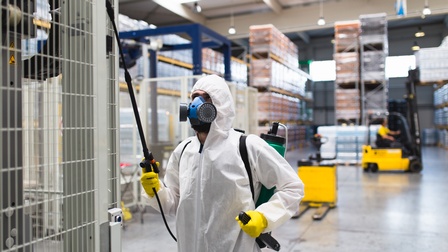 Person in weißem Schutzanzug mit gelben Handschuhen und Atemschutz hält Sprühstab auf Gitterbox in großer Haller, im Hintergrund verschwommen gelber Gabelstapler und befüllte Lagerregale