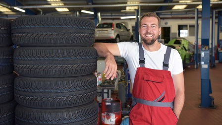 Lächelnde Person in roter Arbeitslatzhose lehnt an Stapel von Autoreifen, im Hintergrund Autos