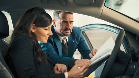 Person hinter Steuer eines PKWs sitzend blickt auf Unterlagen, die andere Person durch Fahrertüre ins Fahrzeuginnere reicht