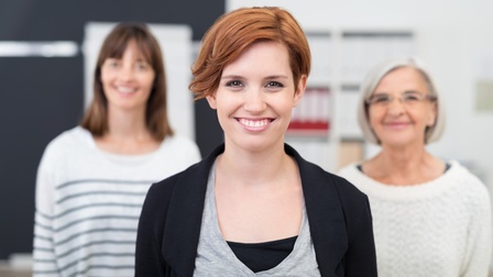 Portrait einer Person mit kurzen rötlichen Haaren blickt freudig in die Kamera, im Hintergrund stehen zwei weitere Personen in der Unschärfe