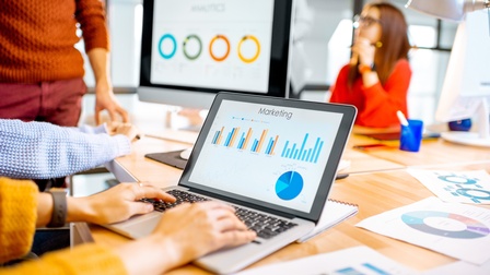 Auf einem großen Tisch steht ein aufgeklappter Laptop, auf dem eine Person tippt. Auf dem Bildschirm sind Diagramme und Statistiken mit der Überschrift Marketing. Am Tisch liegen weitere lose Zettel mit Statistiken