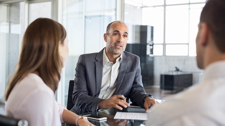 Personen in Businesskleidung sitzen bei einem Besprechungstisch und unterhalten sich, eine Person im Fokus mit Stift in Hand und Dokument vor sich liegend