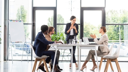 Drei Personen sitzen an einem Tisch, auf dem Gläser und aufgeklappte Laptops sind. Eine Person steht an dem Tisch und ist zur Kamera gewandt. die sitzenden Personen blicken die stehende Person an. Hinter ihnen ist eine große Fensterfront