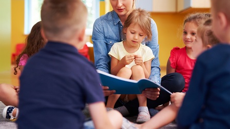 Kinder sitzen um eine Kindergärtnerin, die aus einem Buch vorliest