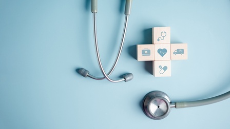 Top-Shot eines Stethoskops sowie von vier Holzwürfeln, die ein Kreuz bilden, auf hellblauem Hintergrund. Auf jedem Würfel ist ein anderes Symbol: ein Herz mit Frequenzlinie, eine Tablette, ein Krankenwagen, ein weiteres Stethoskop und ein Koffer mit Kreuz
