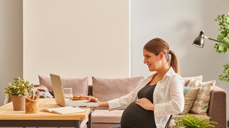 Schwangere Person sitzt freudig bei einem Wohnzimmertisch und arbeitet mit einem Laptop während im Hintergrund eine Couch sowie Pflanzen stehen