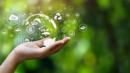 Hand hält eine Visualisierung der Weltkugel mit Icons zum Thema Nachhaltigkeit, Zukunft, ethischer Wirtschaft vor einem grünen Hintergrund mit Bokeh